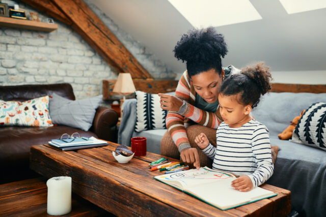 African American mother talks to her small daughter who is drawing on paper at home.