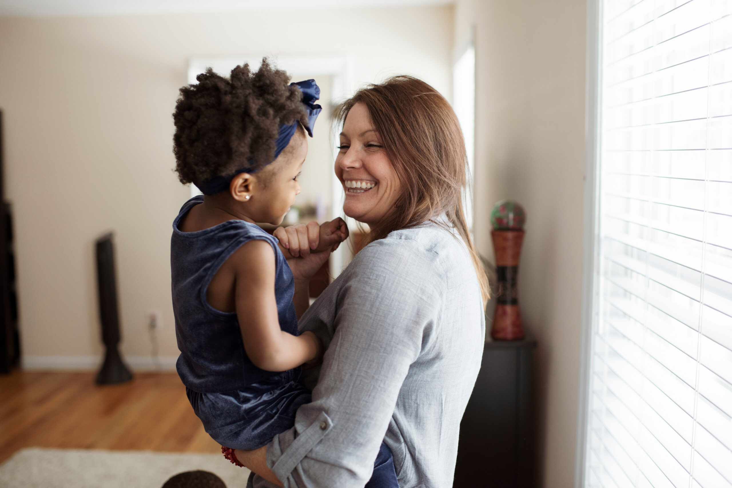 Side view of happy mother carrying cute daughter while standing against window at home
