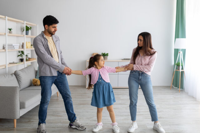 Divorce concept. Arab parents fighting over their child, mad man and woman quarrelling, pulling daughter's hands in different directions, standing in living room interior