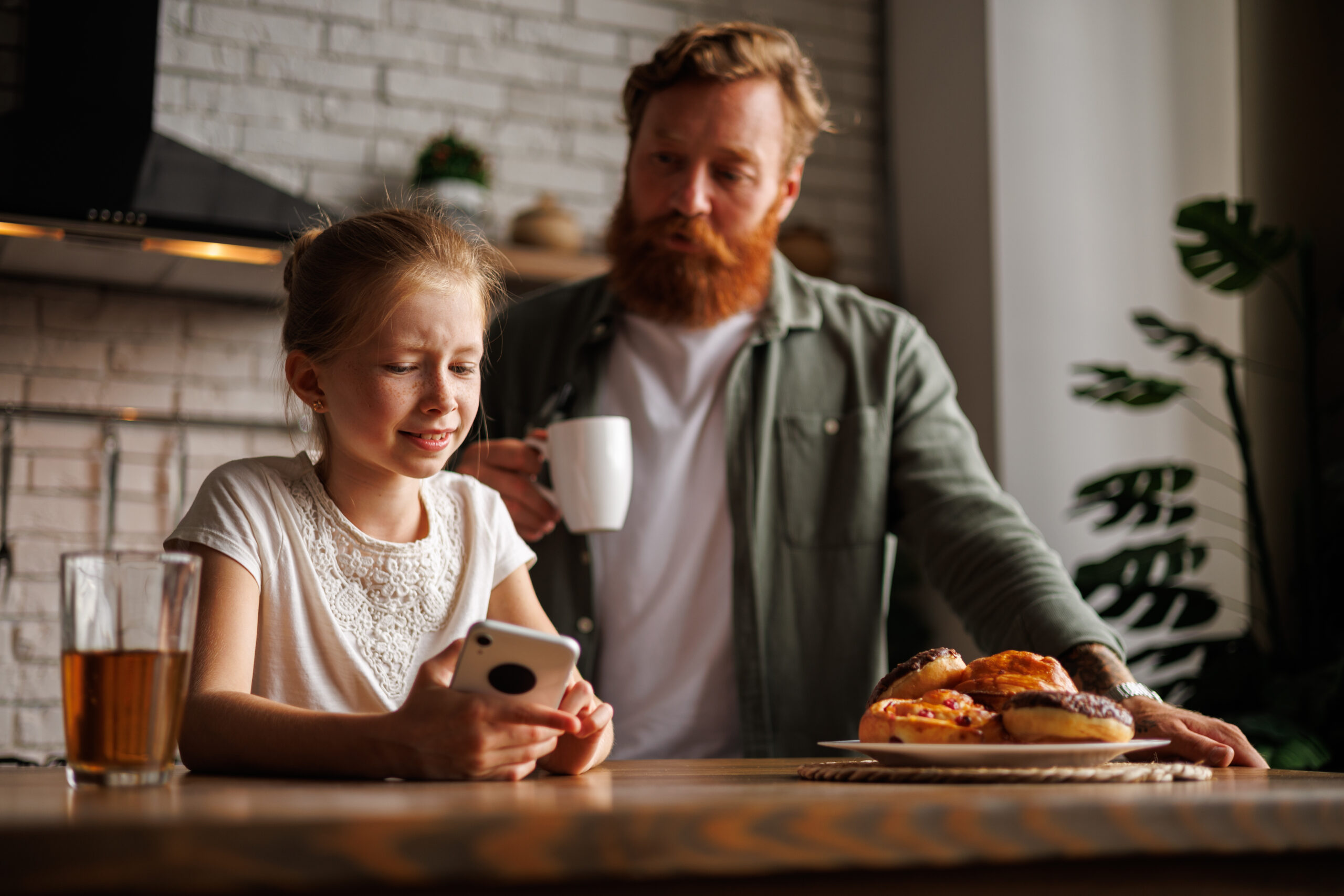 Confused freckled girl showing smartphone to blurred dad with coffee near pastry in kitchen, internet addiction concept