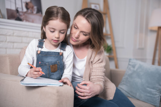 Learning together. Concentrated little girl sitting on the couch with her mother and writing